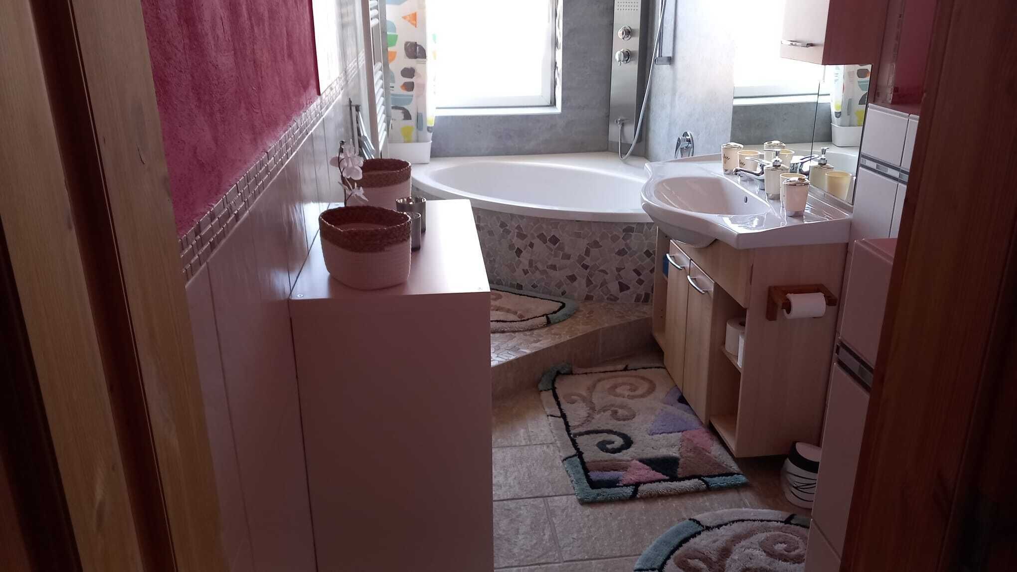 A bathroom with a bathtub, a sink, and a toilet. There are two rugs on the floor and a window providing natural light.