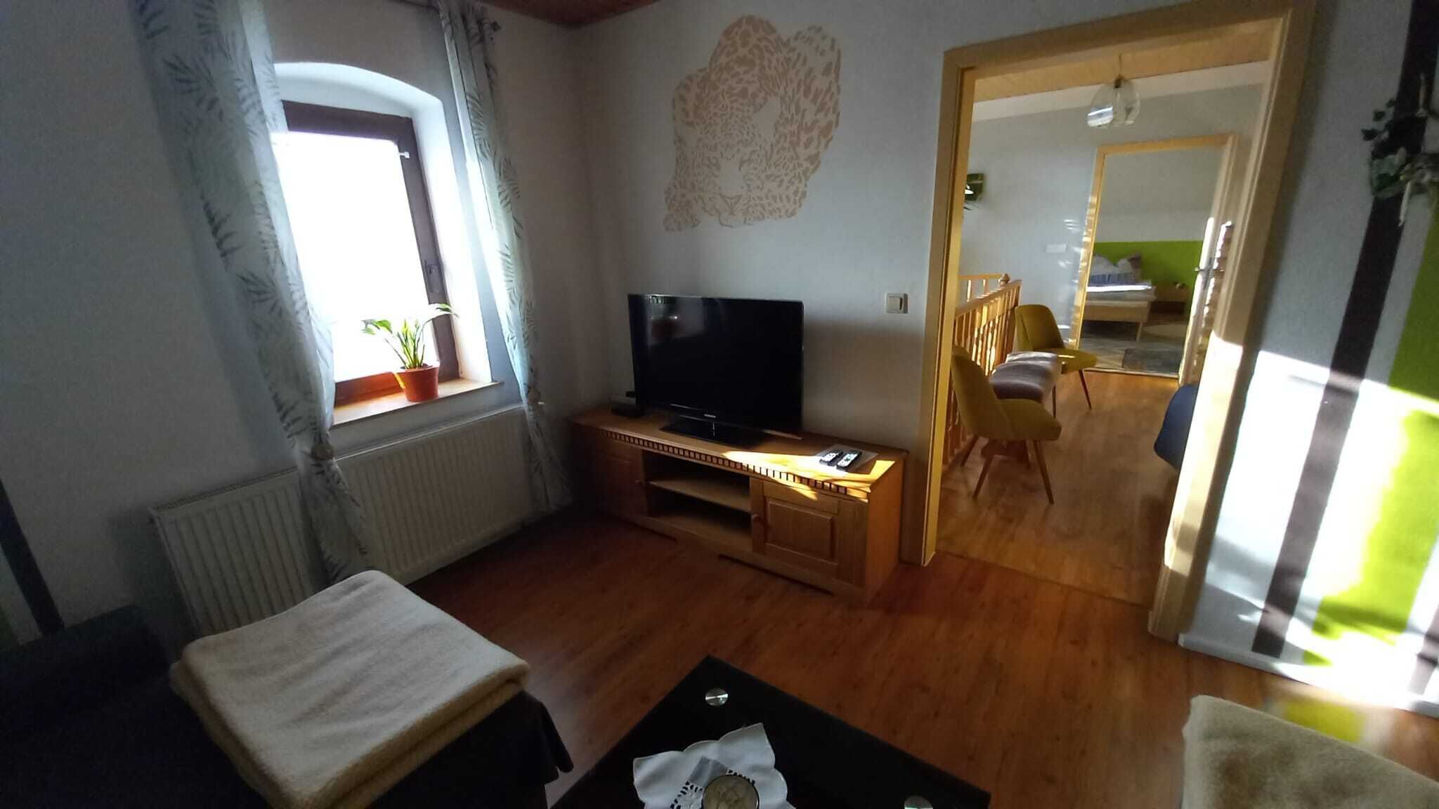 Un salon avec un téléviseur posé sur un meuble en bois, un canapé, une petite table et une fenêtre avec des rideaux.