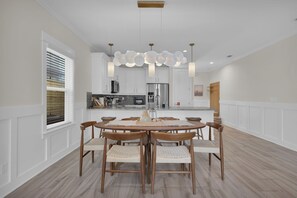 A dining area with a round table, six chairs, pendant lighting, and a kitchen in the background.