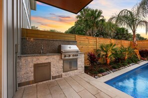 An outdoor kitchen with a grill, built-in oven, and sink, adjacent to a swimming pool with a wooden fence and tropical plants.