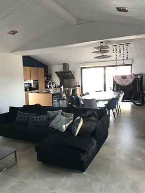 A modern living room with a black sectional sofa, a dining area with a table and chairs, a kitchen with wooden cabinets and a large stove, and a large window providing natural light. - Maison Neuve Idéale Pour les Familles, Proche Océan et Centre Village (Le Bois-Plage-en-Ré)