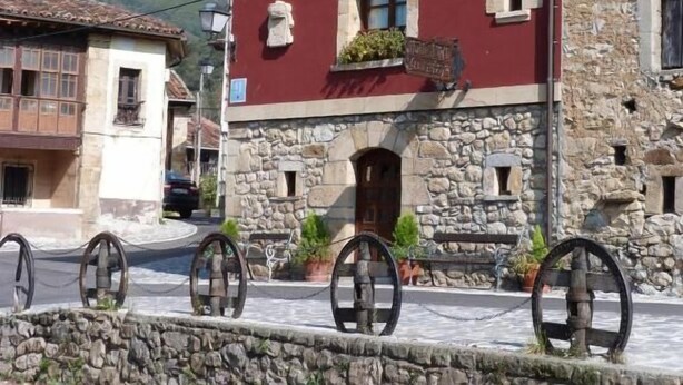 A two-story building with a red facade and stone base, featuring a balcony with potted plants. - Hotel Mestas (Cabrales)
