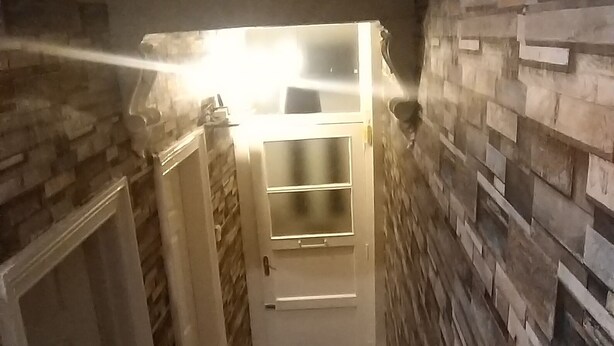 Ein schmaler Flur mit Holzboden, einer schwarzen Matte am Fuß der Treppe und einer Wand mit Steinmuster.