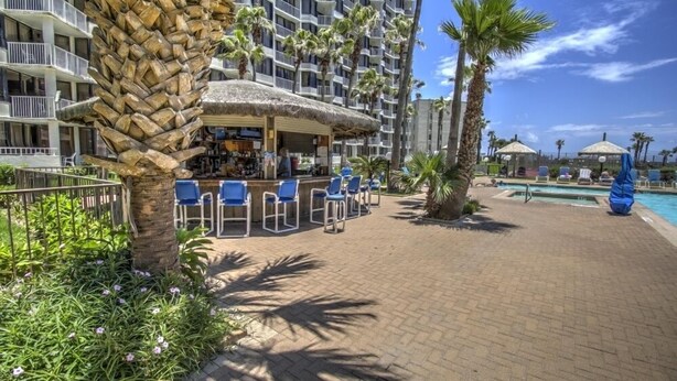 A hotel with a bar area, outdoor seating, a pool, and palm trees.