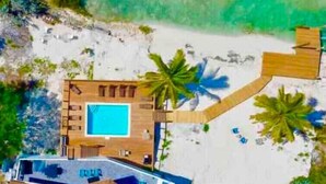 Luchtbeeld van een strandpand met zwembad, meerdere kamers en een strandgedeelte met palmbomen.