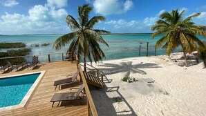 Een houten terras met lounge fauteuils, een zwembad en een zandstrand met palmbomen.