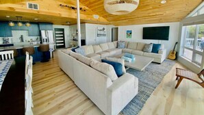 Een ruime woonruimte met een grote L-vormige bank, een salontafel en een eethoek met een lange tafel en stoelen. De kamer heeft houten vloeren, een hoog plafond met zichtbare balken en grote ramen die zorgen voor natuurlijk licht.