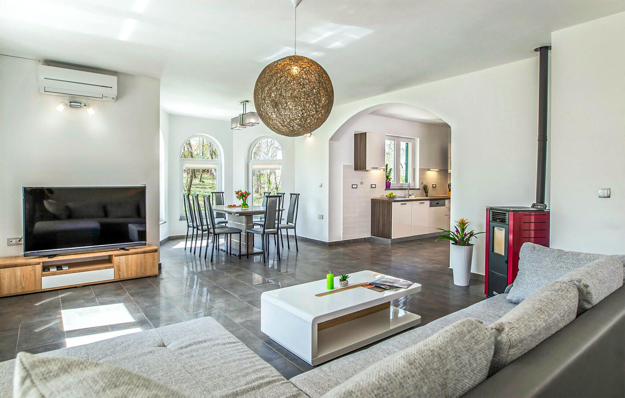 A modern living room with a flat-screen TV, a white coffee table, and a grey sofa. There is a dining area with a table and chairs, and a kitchen with a red cabinet.