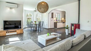 A modern living room with a flat-screen TV, a white coffee table, and a grey sofa. There is a dining area with a table and chairs, and a kitchen with a red cabinet. - Awesome home in Labin (Labin)
