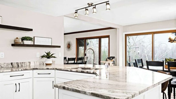 Una cocina moderna con armarios blancos, encimera de mármol y una isla grande. Se ve un comedor con una mesa y sillas a través de un ventanal.