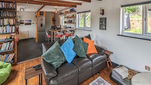 A cozy living room with a leather sofa, a coffee table, and a rug. There is a bookshelf filled with books, a kitchen area with a refrigerator and a dining table, and a window with a view of greenery. - 2 bedroom accommodation in Rye (Rye)