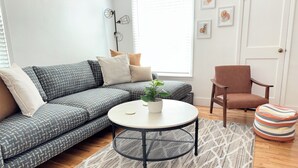 Ein modernes Wohnzimmer mit einem grauen, gemusterten Sofa, einem runden Couchtisch mit einem Topf und Pflanze, einem Holzstuhl und einem gestreiften Hocker.