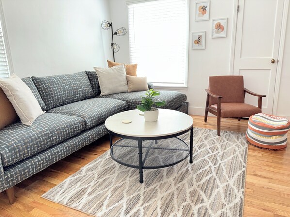 Una sala moderna con un sofá gris estampado, una mesa redonda de centro con una planta en maceta, una silla de madera y un reposapiés a rayas.