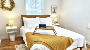 Ein ordentlich eingerichtetes Schlafzimmer mit Bett, Nachttischlampen, einem Nachttisch mit Lampe, einer kleinen Topfpflanze, einem gerahmten Bild an der Wand und einem Fenster mit Jalousien.