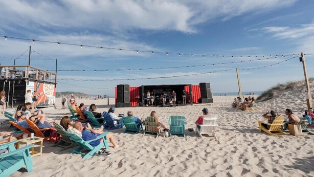 Ein Strand mit sitzenden Personen auf Stühlen, einer Bühne mit roten Vorhängen und Lichterketten darüber.