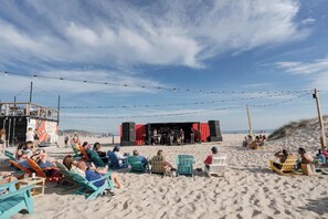 Una playa con gente sentada en sillas, un escenario con cortinas rojas y luces colgantes.