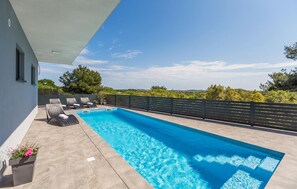 Ein rechteckiger Swimmingpool mit gefliestem Sonnendeck, umgeben von einem schwarzen Zaun. In der Nähe des Pools befinden sich Liegestühle und ein Topfpflanze.