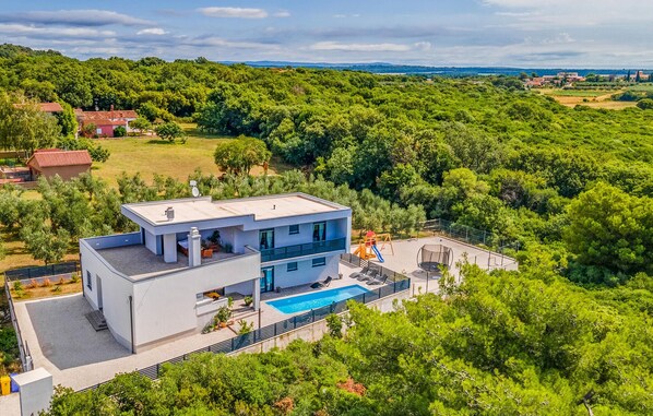 Ein modernes, zweistöckiges Haus mit Pool auf dem Dach, umgeben von üppiger Vegetation und Hügeln im Hintergrund.