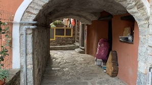 A narrow stone pathway under an arched stone structure leading to a building with a wooden door and a small shelf with items on it. - LA GHIANDA APPARTAMENTO CON TERRAZZO ANIMALI AMMESSI (Cossogno)