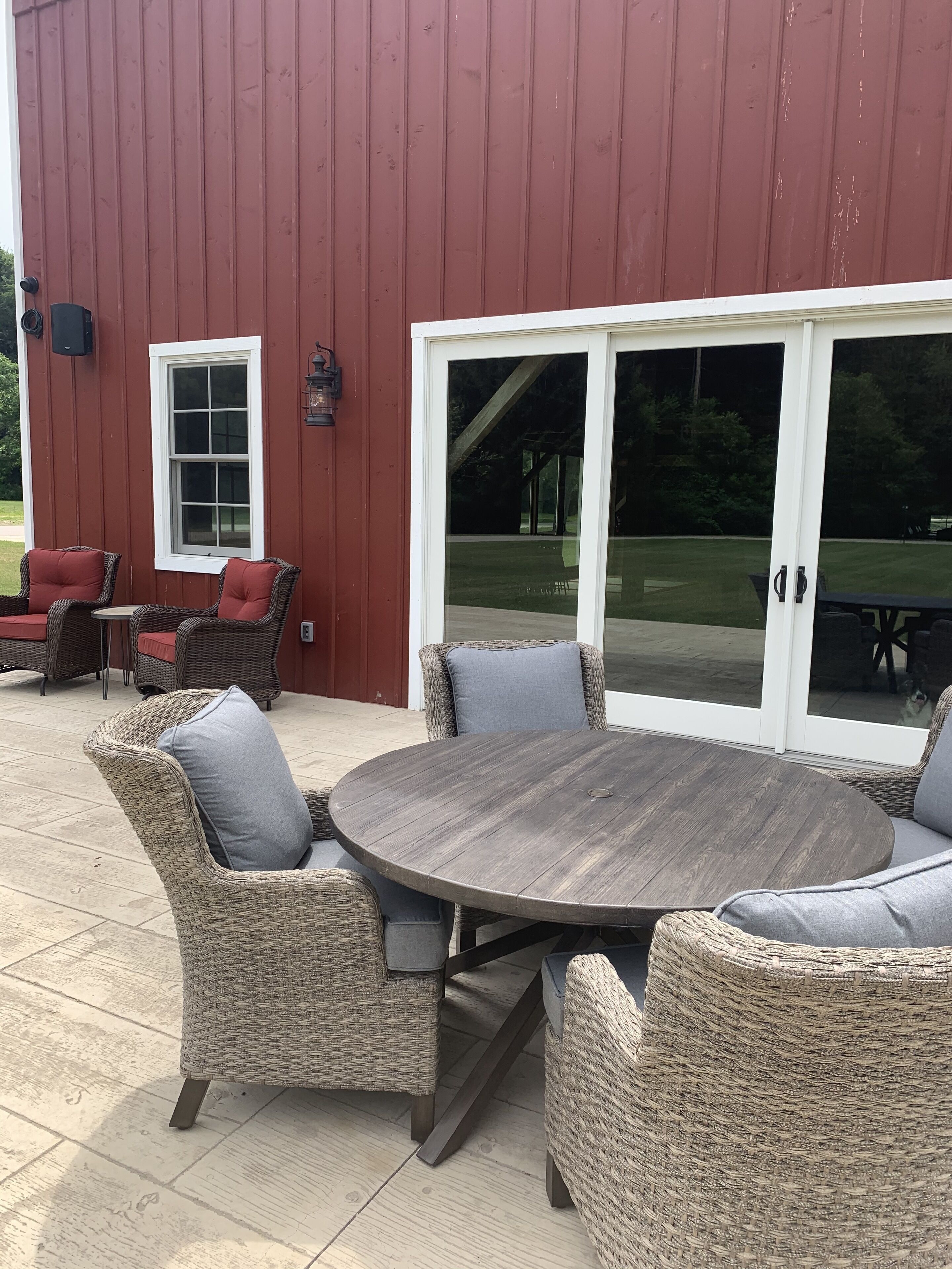 A patio area with wicker furniture and a round table, a red building with a white window, and a sliding glass door leading to a grassy yard.