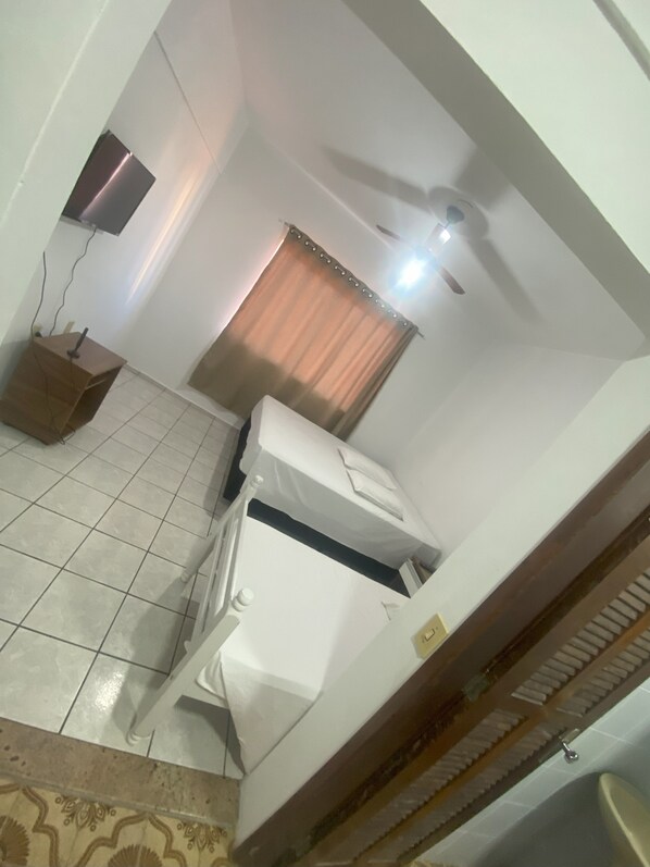 Una habitación pequeña con cama, una mesita y ventilador de techo.