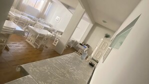 Una habitación con una cama larga y estrecha, una mesita de noche pequeña y una mesita auxiliar. Hay una ventana con cortinas, un estante en la pared con una planta y una puerta con pomo.