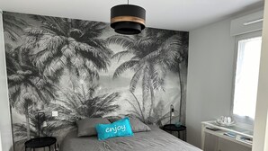 Une chambre avec une grande fresque murale représentant des palmiers, un lit avec une literie grise, un coussin bleu orné du mot « profitez-en », deux tables de chevet noires et une fenêtre avec un rideau blanc.