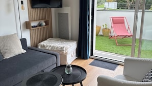 Un salon moderne comprenant un canapé, une table basse, un téléviseur à écran plat fixé sur un panneau en bois, une petite étagère et une porte-fenêtre coulissante donnant sur un balcon où se trouve une chaise rouge.