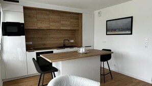 Une cuisine moderne avec des armoires blanches, un plan de travail en bois et un micro-ondes noir. On y trouve un îlot central en bois, doté d’un évier et d’un vase blanc. Deux tabourets de bar noirs sont placés à côté de l’îlot. Une chaise blanche se trouve au premier plan.
