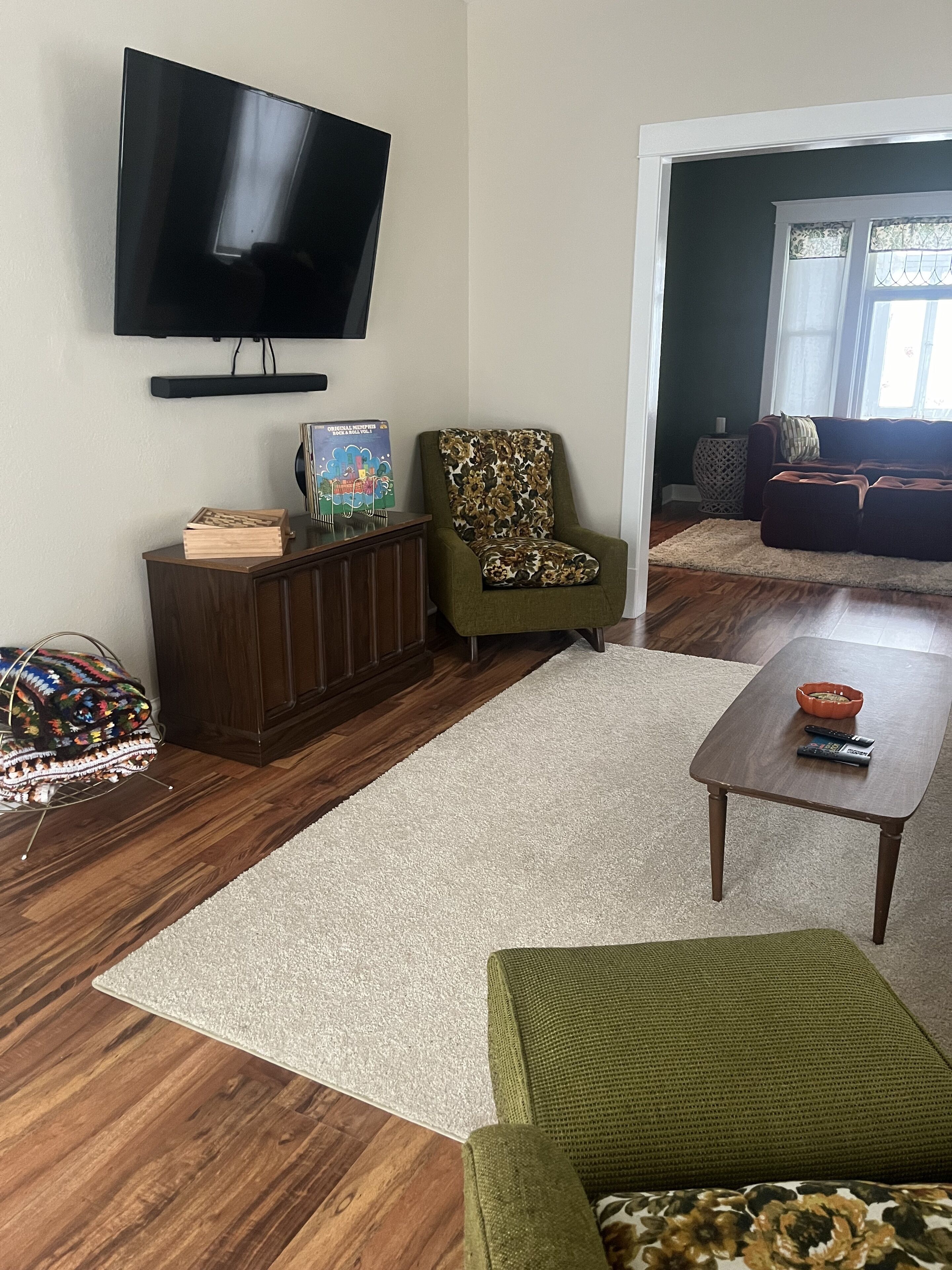 A living room with a flat-screen TV mounted on the wall, a wooden cabinet, a patterned armchair, a green sofa, and a coffee table.