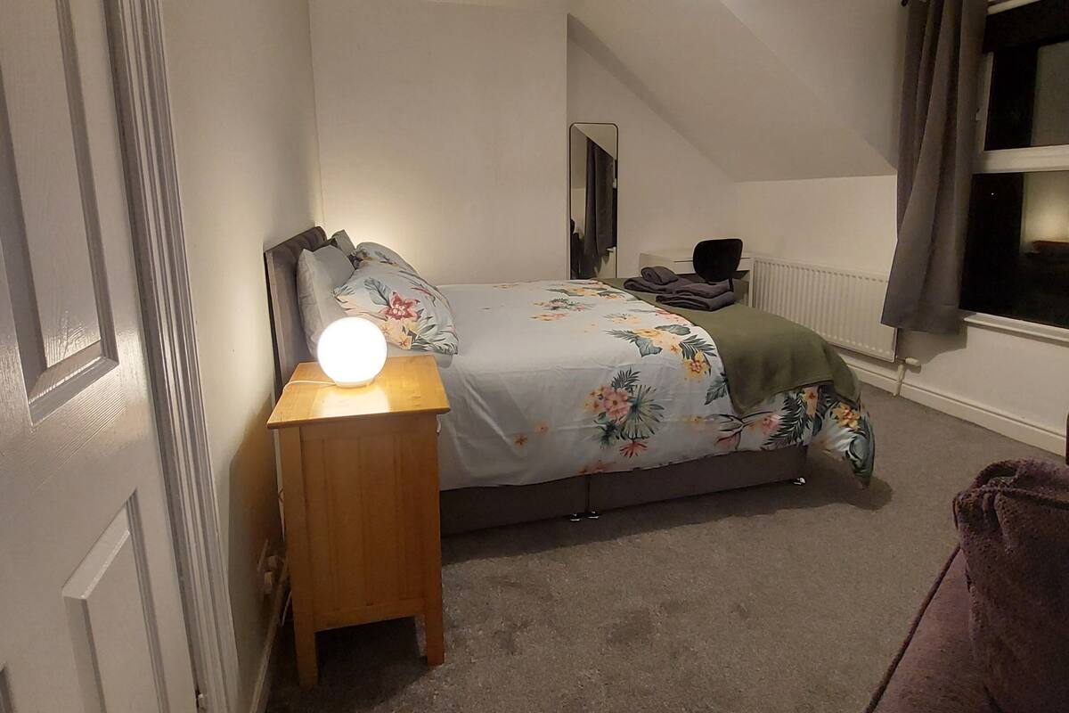 Un dormitorio con cama, lámpara de noche, espejo, silla y ventana con cortinas.