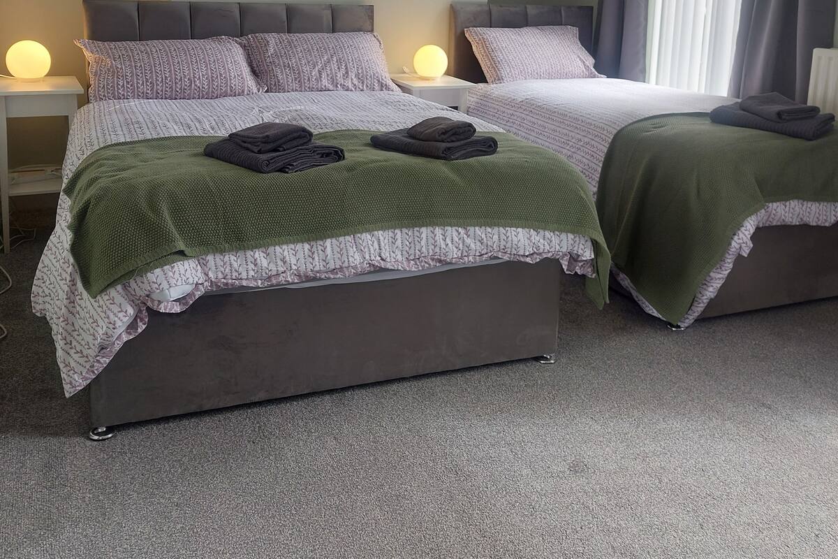 Habitación de hotel con dos camas, cada una con cabecera y almohadas, una colcha verde en una cama y una blanca en la otra.