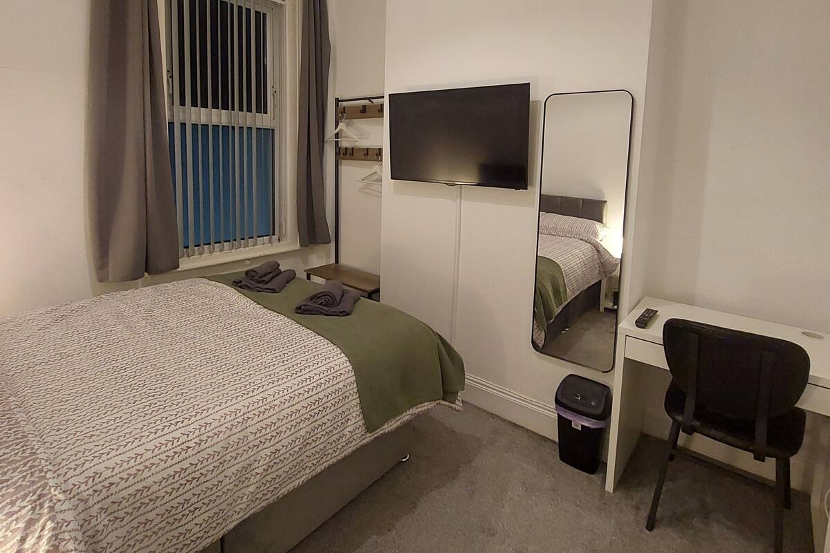 Un dormitorio con una cama, un escritorio, una silla, un televisor y una ventana con cortinas.