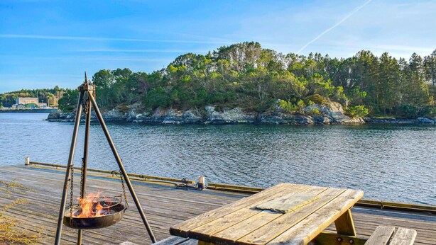 Un muelle de madera con una parrilla y una mesa de picnic, con vistas a una masa de agua tranquila y una orilla boscosa.