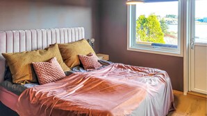 Un dormitorio con una cama, almohadas, una mesita de noche y una ventana con vistas a árboles y edificios.