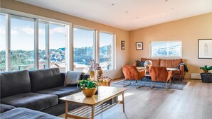 Una sala de estar moderna con sofá, mesa de centro y sillas. Amplias ventanas ofrecen vistas de un paisaje urbano.