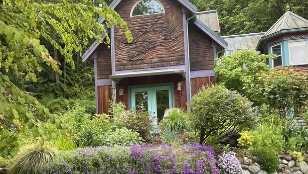 A wooden house with a green door, surrounded by lush greenery and purple flowers. - The Loft at Reverie Gardens (Sequim)