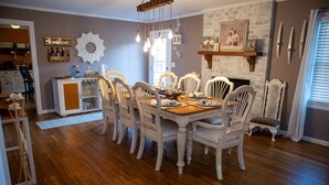 Une salle à manger avec une table en bois et des chaises blanches, une cheminée et un coin cuisine équipé d’un réfrigérateur et de placards.