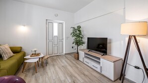 Una sala de estar moderna con un sofá, una mesita, un televisor sobre un mueble de madera, una lámpara y una planta en maceta.