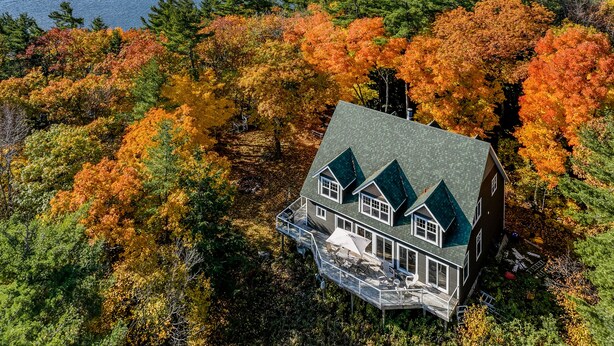 A large house with a green roof and a deck, surrounded by autumn-colored trees and a lake.