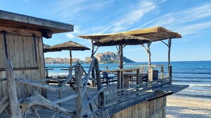 A wooden beachside structure with thatched roofs, a wooden deck, and a view of the sea and a distant city. - Petite Maison Dans Résidence 3 Etoiles Avec Piscine (Belgodère)