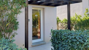 A modern building with a covered entrance, a small garden area with green plants, and a clear sky. - Petite Maison Dans Résidence 3 Etoiles Avec Piscine (Belgodère)