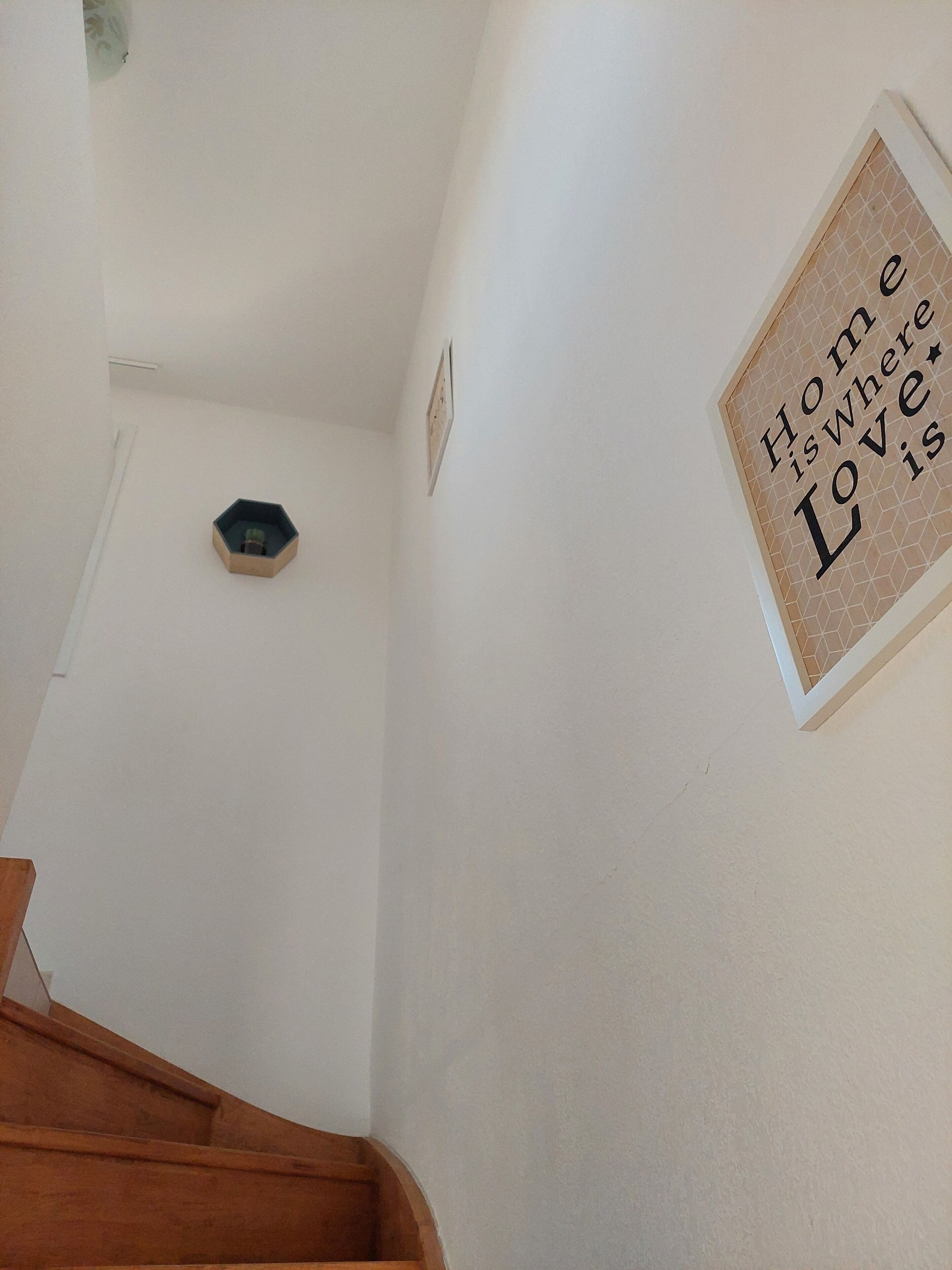 Un escalier avec une rampe en bois menant à un palier. Une plaque décorative accrochée au mur porte l’inscription : « Le foyer, c’est là où se trouve l’amour ».