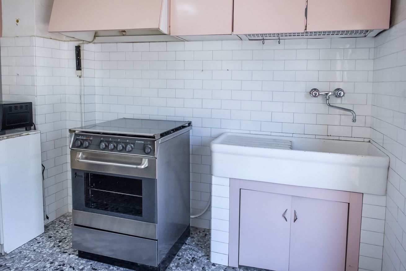 A kitchen with a stove, oven, and sink, white tiled walls, and pink cabinets.