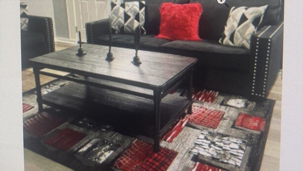Een woonkamer met een zwarte bank, een tapijt met rood-wit patroon en een zwarte salontafel.