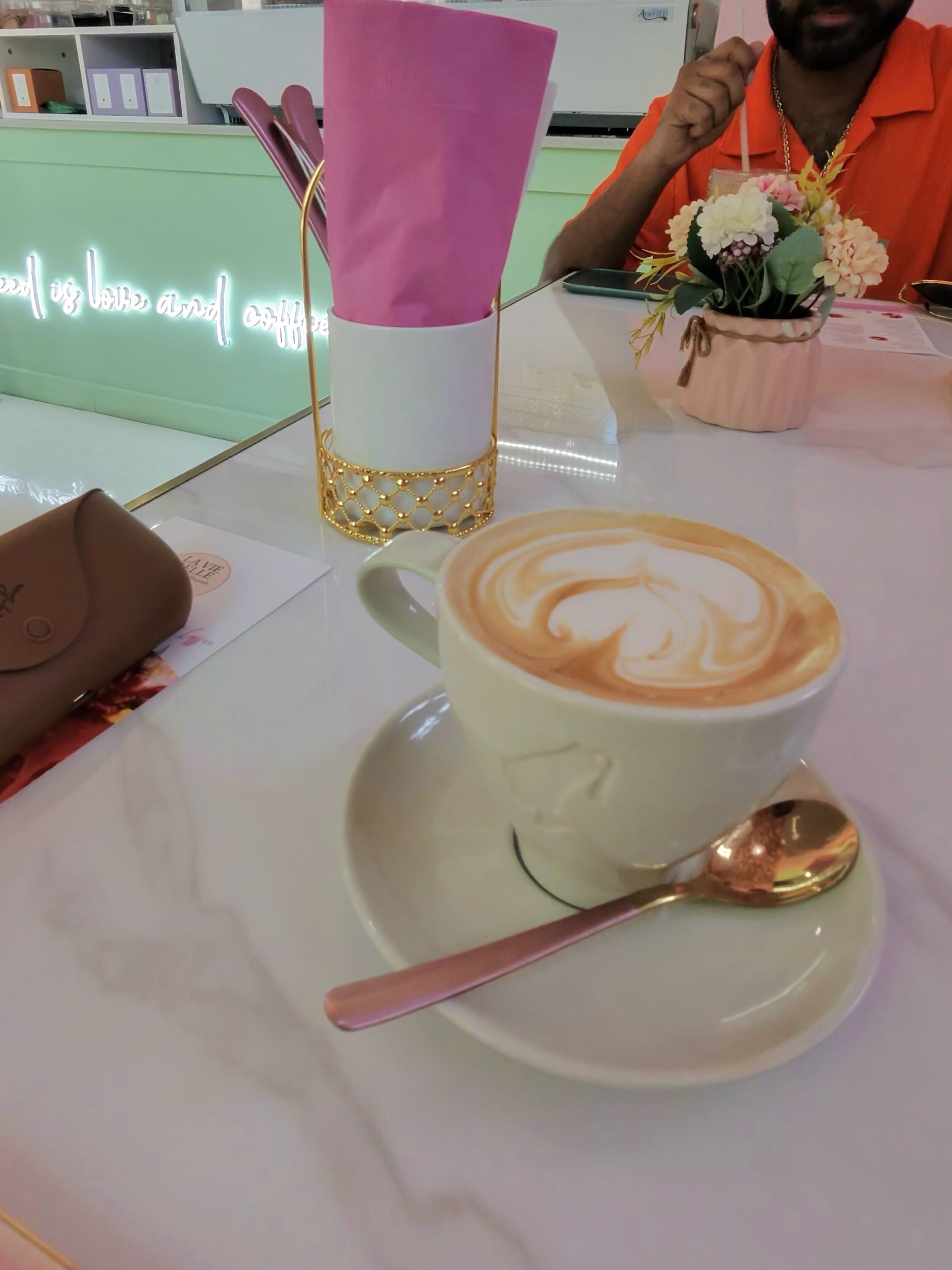 Una taza de café con arte de latte sobre un plato, una cuchara, un recipiente rosa para servilletas y un pequeño jarrón con flores sobre una mesa.