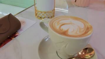 Una taza de café con arte de latte sobre un plato, una cuchara, un recipiente rosa para servilletas y un pequeño jarrón con flores sobre una mesa.