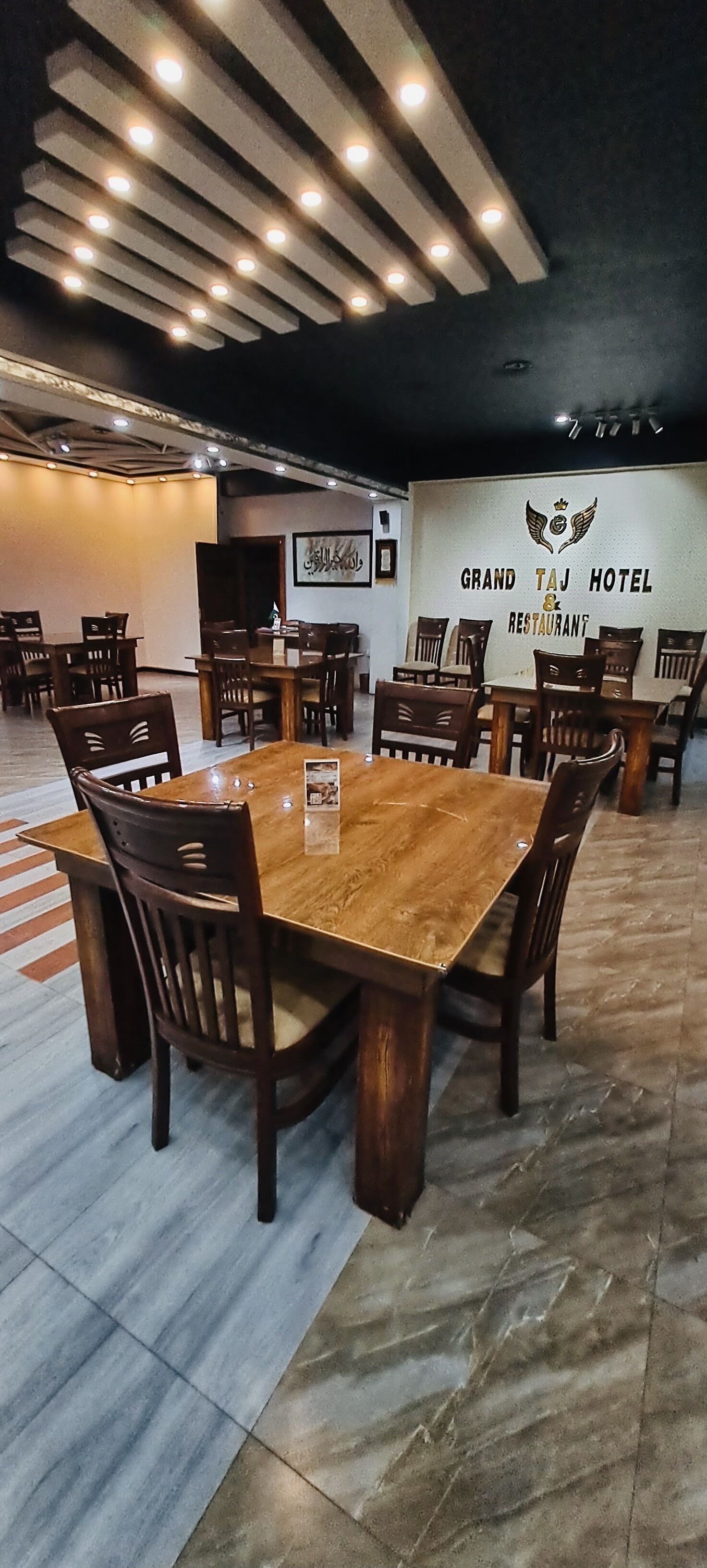 Un amplio comedor con varias mesas y sillas de madera, un letrero que indica Grand Taj Hotel & Restaurant, y un techo moderno con iluminación empotrada.