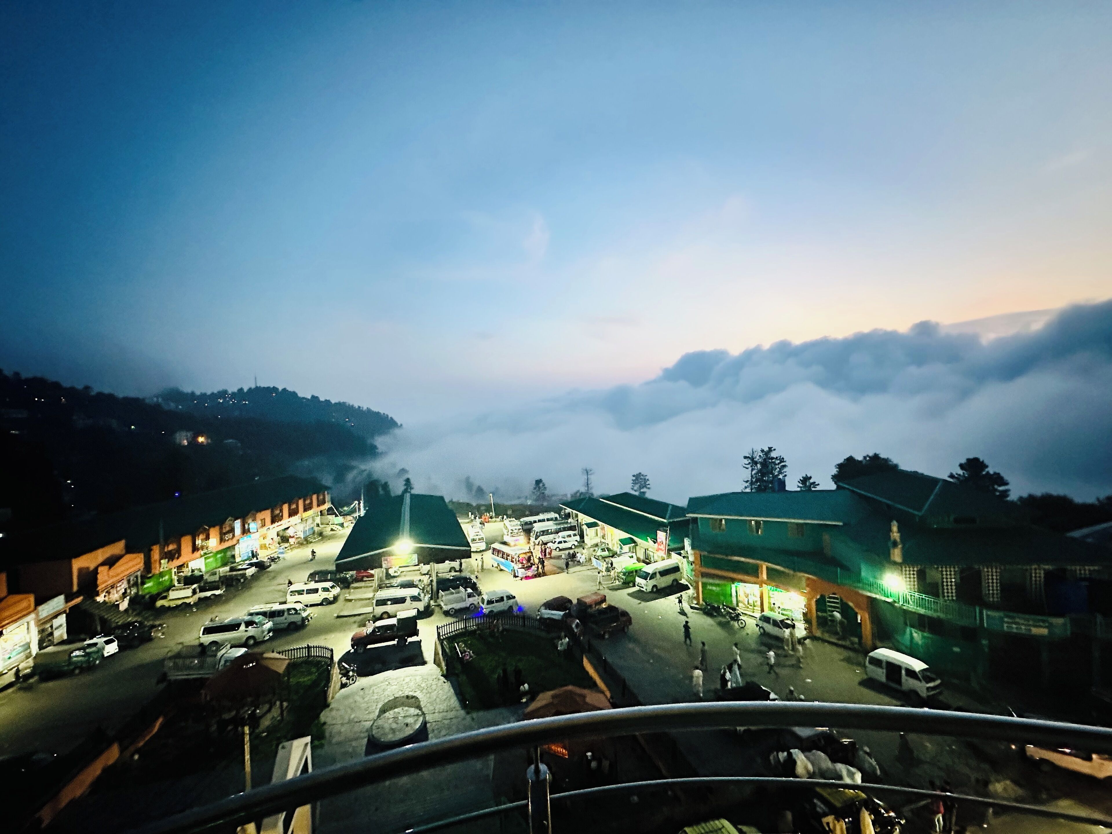 A town square at dusk with buildings, vehicles, and people.