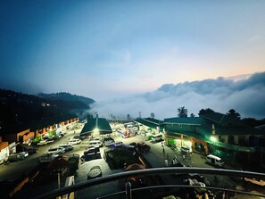 Une place de ville au crépuscule, avec des bâtiments, des véhicules et des passants.
